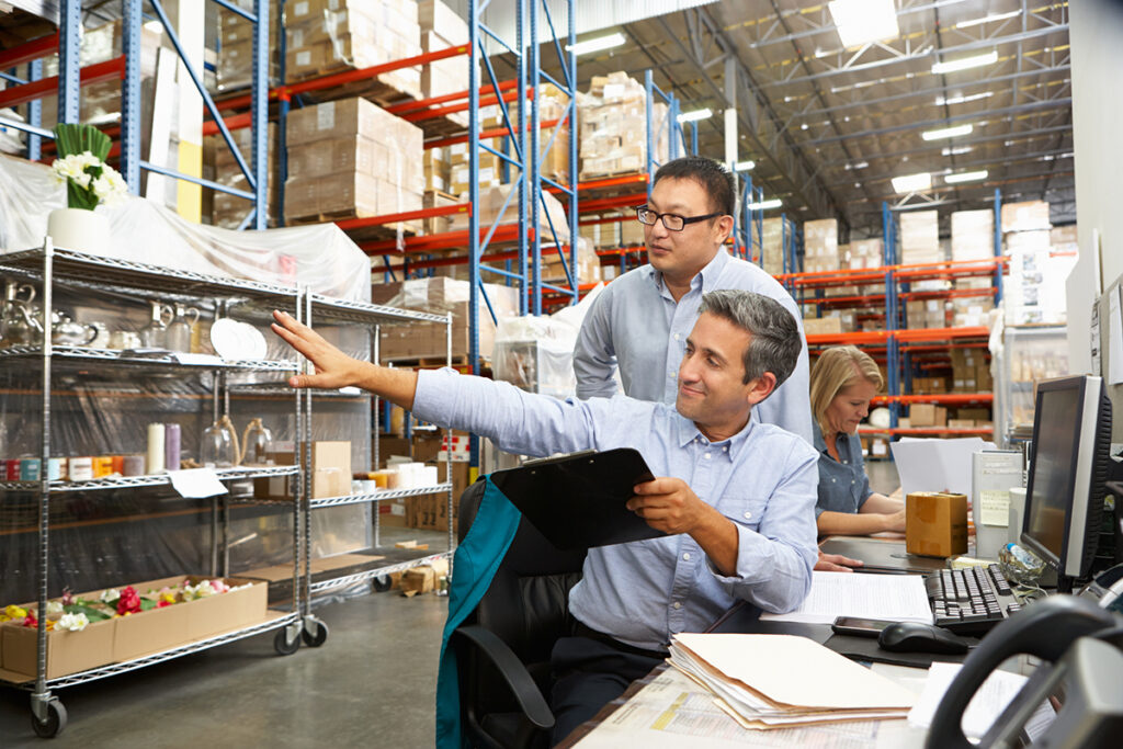 Business Colleagues Working At Desk In Warehouse Holding Clipboard